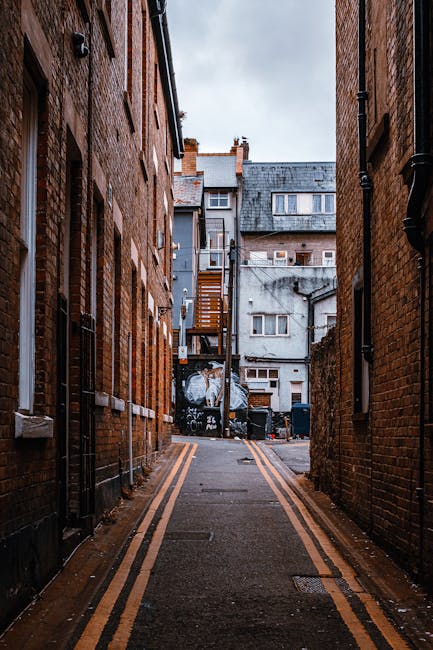 A narrow alleyway between two brick buildings with double yellow lines on the asphalt surface, leading towards a residential area at the end. Visible at the far end are multi-storey houses with various window styles and external features such as chimneys, balconies, and external staircases, some with wooden railings. The alley is outdoors under cloudy sky lighting, with black drainage or utility pipes running along the building walls. In the distance, there are several black trash bins and a large recycling container. This setting illustrates the type of confined access environment that Man and Van Teddington may encounter during house removal or furniture transport operations, especially when navigating narrow spaces before loading items onto a van for home relocation or packing and moving processes.
