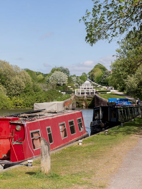 A narrowboat with a red exterior, partially covered with a grey tarp, is moored along the bank of a canal near a grassy towpath. Adjacent to it, a black narrowboat is also moored, with a blue storage container on its roof. The scene is set on a bright, clear day with a blue sky and a few clouds, surrounded by trees with lush green foliage. In the background, there is an ornate white footbridge crossing the canal, and beyond it, a tree-lined walkway with blooming trees, indicating a park environment. The image captures a peaceful setting suitable for planning a home relocation involving furniture transport and loading activities, with Man and Van Teddington occasionally involved in house removals or moving logistics.