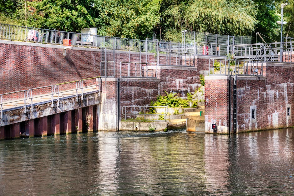 View of the brick and concrete retaining wall along the edge of a waterway, with metal railings, stairs, and platforms for access. The wall shows signs of weathering with some discoloration and moss growth. There are plants growing at the riverbank near the steps. The scene is outdoors during daytime, with the water reflecting the structures and greenery above, and trees with dense foliage in the background. This setting is typical for a riverside location where furniture and boxes might be loaded or unloaded during a home relocation or furniture transport process, as handled by companies like Man and Van Teddington. The environment indicates a well-maintained area suitable for loading and unloading during moving services, aligning with the context of packing and loading for a house removal from Bushy Park to Teddington Lock.