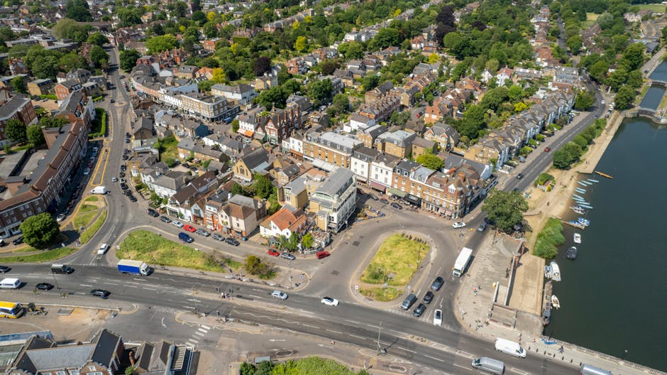 A street view on Ferry Road TW11 showing a row of multi-storey terraced houses with brick facades and gabled roofs, some with small balconies. The pavement is lined with pedestrians and shopfronts, in