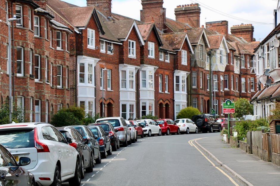 A residential street with a row of traditional red-brick terraced houses featuring bay windows and pitched roofs, set under a partly cloudy sky. Parked along the curb are numerous cars, including hatchbacks, sedans, and small SUVs, occupying the designated parking spaces. The street curves gently to the right, with a narrow sidewalk on the right side bordered by low fencing and small gardens. A street sign is visible, indicating parking regulations enforced by Richmond Council, which may affect home relocation and furniture transport processes. In the foreground, the pavement and roadside are clean, and the parked vehicles are aligned neatly. This scene captures an urban residential area ideal for house removals, where careful planning of loading and unloading areas is crucial to comply with local parking rules, particularly when scheduling moving and packing activities with a professional service like Man and Van Teddington.