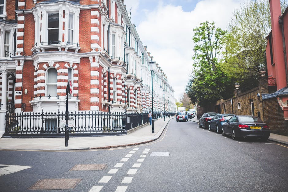A street scene in Teddington showing a row of parked cars along the curb adjacent to residential buildings with Victorian-style architecture featuring red brick facades, white window frames, and decorative elements. The buildings are separated from the pavement by black wrought-iron fences, with some access points leading to doorways. The street includes a designated loading zone marked by white dashed lines on the asphalt, indicating areas for loading and unloading activities related to house removals. There are a few pedestrians visible walking along the pavement. In the background, a white moving truck is parked further down the street, with a man in casual clothing engaged in loading furniture or cardboard boxes onto it, assisted by a dolly or hand truck. The scene is well-lit with natural daylight, and lush green trees line the sidewalk on the right side of the street, contributing to the residential atmosphere. This setting illustrates the urban environment where companies like Man and Van Teddington coordinate home relocation and furniture transport, considering local parking rules affecting removals in the area.