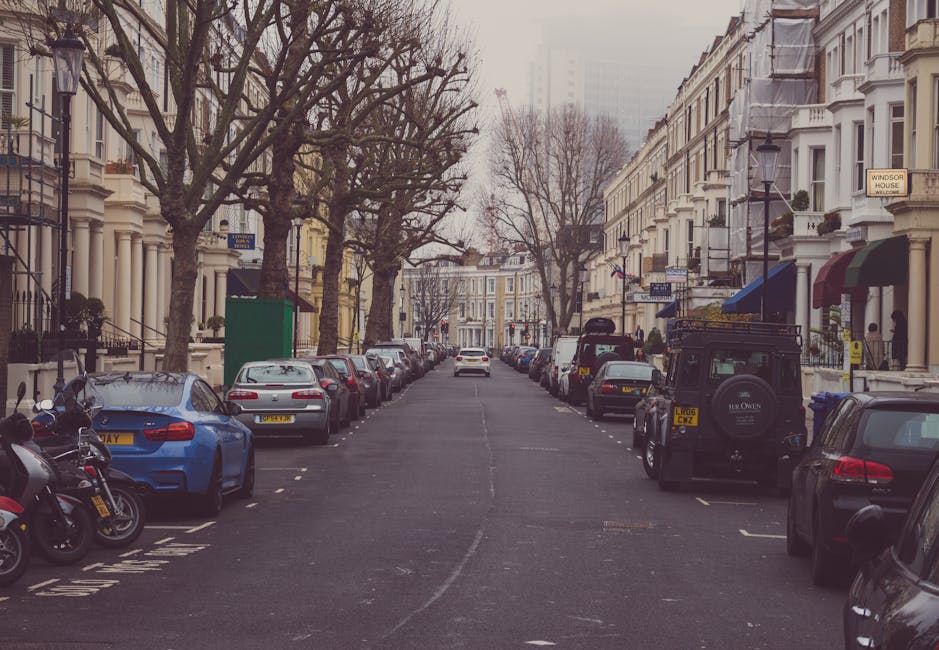 A residential street with a row of traditional red-brick terraced houses featuring bay windows and pitched roofs, set under a partly cloudy sky. Parked along the curb are numerous cars, including hatchbacks, sedans, and small SUVs, occupying the designated parking spaces. The street curves gently to the right, with a narrow sidewalk on the right side bordered by low fencing and small gardens. A street sign is visible, indicating parking regulations enforced by Richmond Council, which may affect home relocation and furniture transport processes. In the foreground, the pavement and roadside are clean, and the parked vehicles are aligned neatly. This scene captures an urban residential area ideal for house removals, where careful planning of loading and unloading areas is crucial to comply with local parking rules, particularly when scheduling moving and packing activities with a professional service like Man and Van Teddington.