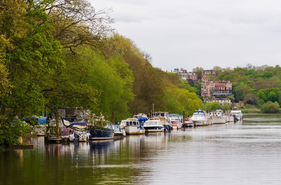A row of various motorboats and yachts docked along the riverbank, with some boats covered in protective tarps or plastic wrapping, while others are uncovered exposing seating and cabin areas. The vessels are tied to the quayside, close to the edge of the water, with metal posts and mooring lines securing them. Large green-leaved trees extend over part of the boats, providing partial shade, and their branches reach towards the water. In the background, there are residential buildings on a hillside, some with exposed brick and ornate architectural details, surrounded by lush greenery. The calm river reflects the boats and trees, creating a peaceful scene suitable for house removal and moving services that involve transporting boats or relocating along the waterfront. The atmosphere is overcast, with soft lighting enhancing the natural colours and textures of the scene, illustrating a typical riverside setting in Teddington. Man and Van Teddington may handle such waterfront transportation and home relocation logistics for clients in the area.