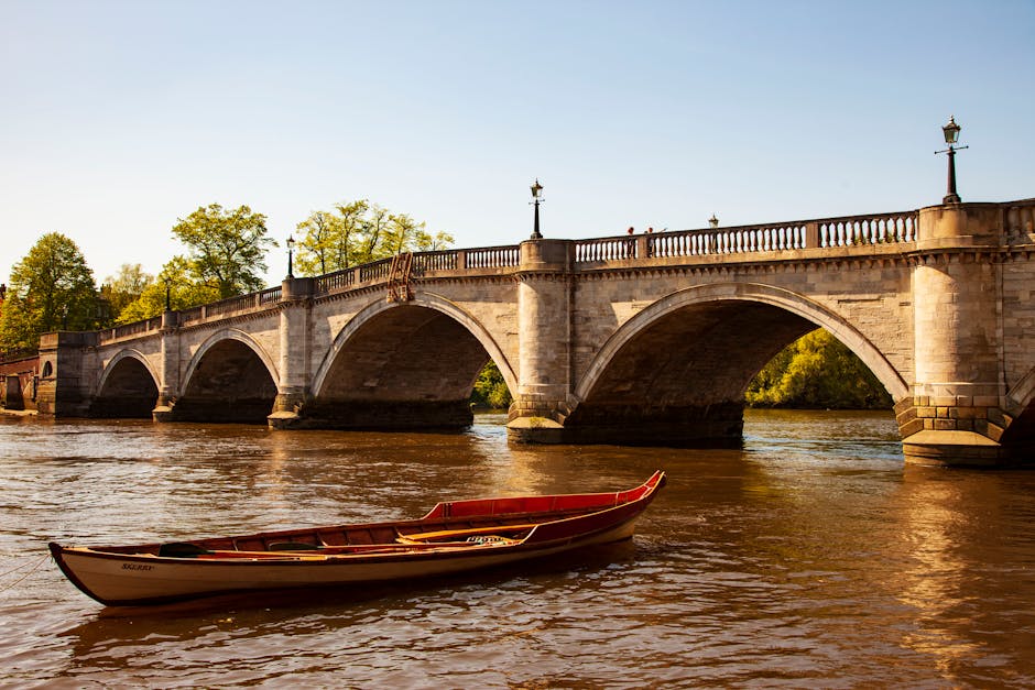 A historic stone bridge with multiple arches spanning across a calm river, featuring decorative street lamps along its balustrade. The bridge is illuminated by warm sunlight, casting shadows on the water's surface. In the foreground, a small wooden boat with a red interior and a white exterior floats close to the riverbank, where a man and woman are seen loading a cardboard box wrapped in plastic and secured with straps onto a wheeled trolley. The scene takes place outdoors on a clear day, with lush green trees visible behind the bridge. The setting suggests a home relocation process involving furniture transport and packing, with the scene captured to illustrate the logistics of moving items from a residence during a house removal through narrow bridge access or waterfront transfer, highlighting the careful handling of packing materials and the use of appropriate equipment. This image aligns with services provided by Man and Van Teddington in accordance with Richmond upon Thames Council rules for Teddington removals.