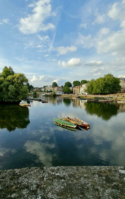 A row of various motorboats and yachts docked along the riverbank, with some boats covered in protective tarps or plastic wrapping, while others are uncovered exposing seating and cabin areas. The vessels are tied to the quayside, close to the edge of the water, with metal posts and mooring lines securing them. Large green-leaved trees extend over part of the boats, providing partial shade, and their branches reach towards the water. In the background, there are residential buildings on a hillside, some with exposed brick and ornate architectural details, surrounded by lush greenery. The calm river reflects the boats and trees, creating a peaceful scene suitable for house removal and moving services that involve transporting boats or relocating along the waterfront. The atmosphere is overcast, with soft lighting enhancing the natural colours and textures of the scene, illustrating a typical riverside setting in Teddington. Man and Van Teddington may handle such waterfront transportation and home relocation logistics for clients in the area.