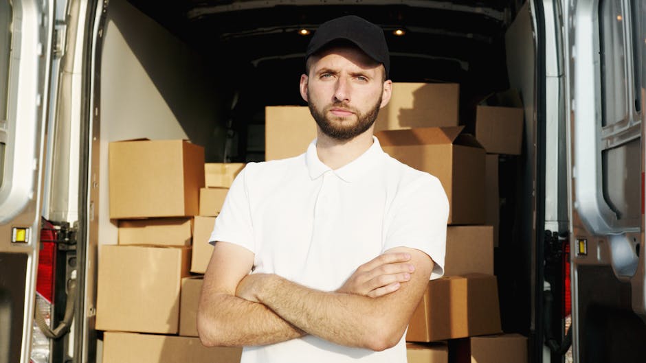 A young man with a serious expression stands inside the open rear of a moving van, which is loaded with multiple cardboard boxes of various sizes, some wrapped in plastic and others in plain cardboard. The man, wearing a black cap and a white polo shirt, has his arms crossed and looks directly at the camera. The moving van is parked on a paved surface, with the interior visible showing the packed boxes arranged for home relocation or furniture transport. The scene depicts the loading process associated with packing and moving services, emphasizing the logistical aspects of house removals. The environment is well-lit, with natural daylight illuminating the interior and the man. This image is relevant to urgent, same-day removal services offered by companies like Man and Van Teddington, highlighting the preparation phase of a swift house move in Teddington, focused on efficient packing and transport.