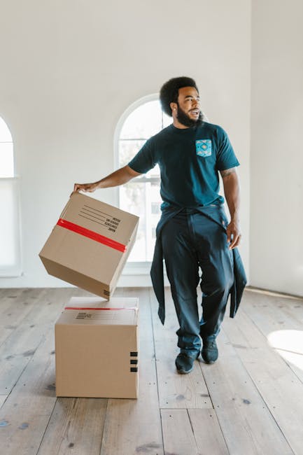 A man with dark curly hair and a beard is inside a bright, spacious room with white walls and a large arched window behind him, allowing natural light to fill the space. He is wearing a dark blue t-shirt with a logo on the chest, dark trousers, and has a piece of clothing tied around his waist. He is holding a cardboard box that is sealed with red packing tape, lifting it from two similarly sealed boxes on the wooden floor. The boxes are standard cardboard cartons used for packing belongings during a house move. The room appears to be part of a home undergoing relocation, with no furniture visible, and the focus is on the man preparing to move or load the boxes. The environment suggests a professional or private house transfer service, typical of furniture transport and packing operations involved in house removals, supported by the presence of the boxes and the interior setting.