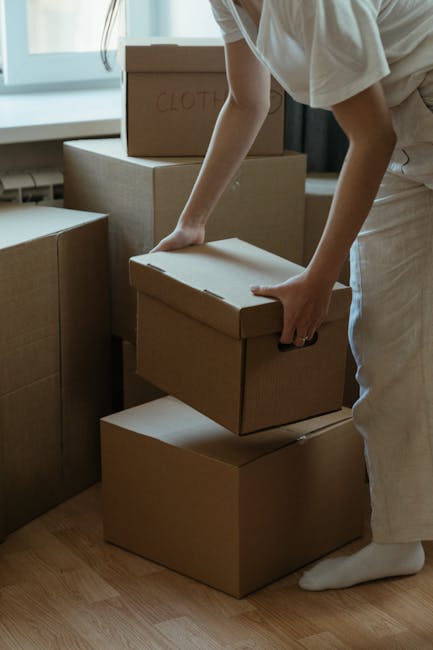 A young man with a serious expression stands inside the open rear of a moving van, which is loaded with multiple cardboard boxes of various sizes, some wrapped in plastic and others in plain cardboard. The man, wearing a black cap and a white polo shirt, has his arms crossed and looks directly at the camera. The moving van is parked on a paved surface, with the interior visible showing the packed boxes arranged for home relocation or furniture transport. The scene depicts the loading process associated with packing and moving services, emphasizing the logistical aspects of house removals. The environment is well-lit, with natural daylight illuminating the interior and the man. This image is relevant to urgent, same-day removal services offered by companies like Man and Van Teddington, highlighting the preparation phase of a swift house move in Teddington, focused on efficient packing and transport.