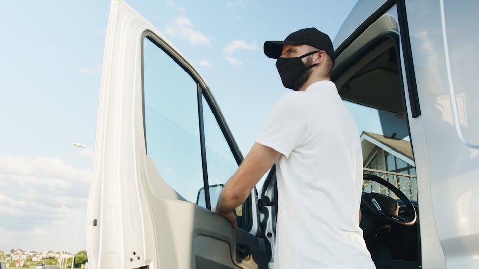 A man wearing a black face mask, black cap, and white t-shirt is standing at the open door of a white moving van, preparing for a home relocation. He is gripping the door handle with both hands, appearing to be either entering or exiting the vehicle. Inside the van, visible packing materials include cardboard boxes and plastic-wrapped items, stacked securely on the vehicle’s floor, which indicates a packing and moving process. The scene is set outdoors on a clear day with a blue sky and scattered clouds, and a residential area with houses and streetlights is faintly visible in the background. The setting captures a typical furniture transport or loading process, with the focus on efficient packing and logistics managed by Man and Van Teddington, involved in professional removals services. The open van door and the positioning of the man suggest the loading or unloading phase of a house removal operation, highlighting the importance of careful packing and transport during the move.