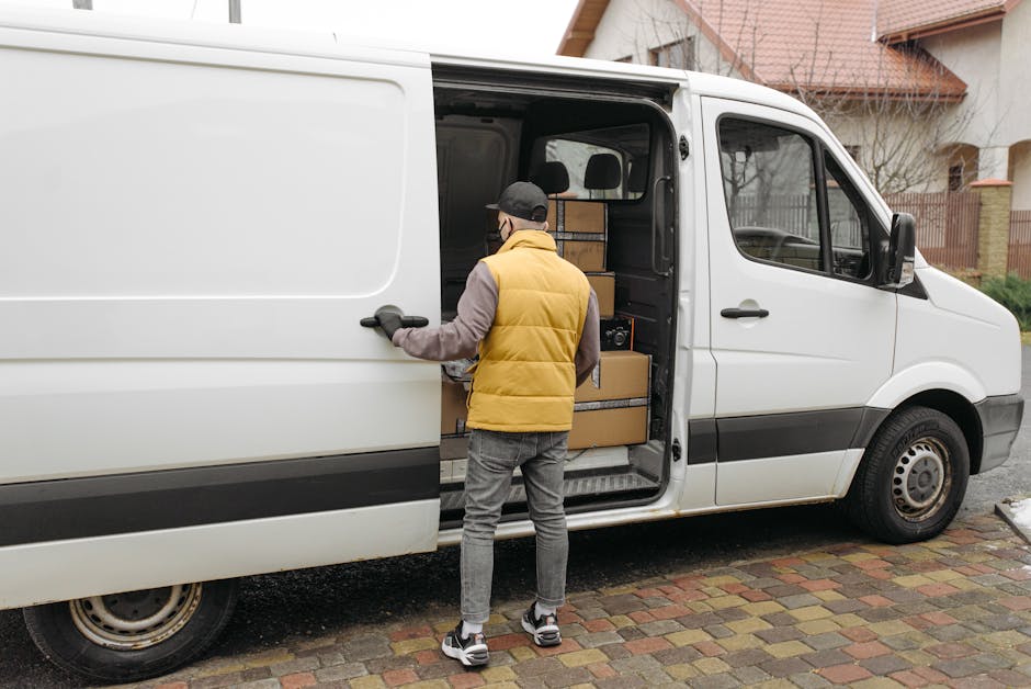 A man wearing a grey cap, brown trousers, grey shoes, and a yellow padded vest is loading a white panel van with cardboard moving boxes of various sizes, some wrapped in black tape. The van's sliding door is open, revealing more boxes stacked inside on a metal platform. The scene is set outside a residential property with a brick-paved driveway and a neighboring house with brown roofing and fencing visible in the background. The process appears to be part of a home relocation, with the man carefully handling the boxes, supported by equipment such as straps or trolleys, illustrating furniture transport and packing and moving activities typical of house removals in Teddington.