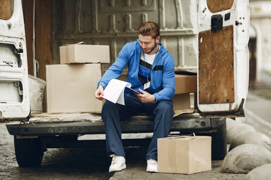 A man wearing a black face mask, black cap, and white t-shirt is standing at the open door of a white moving van, preparing for a home relocation. He is gripping the door handle with both hands, appearing to be either entering or exiting the vehicle. Inside the van, visible packing materials include cardboard boxes and plastic-wrapped items, stacked securely on the vehicle’s floor, which indicates a packing and moving process. The scene is set outdoors on a clear day with a blue sky and scattered clouds, and a residential area with houses and streetlights is faintly visible in the background. The setting captures a typical furniture transport or loading process, with the focus on efficient packing and logistics managed by Man and Van Teddington, involved in professional removals services. The open van door and the positioning of the man suggest the loading or unloading phase of a house removal operation, highlighting the importance of careful packing and transport during the move.
