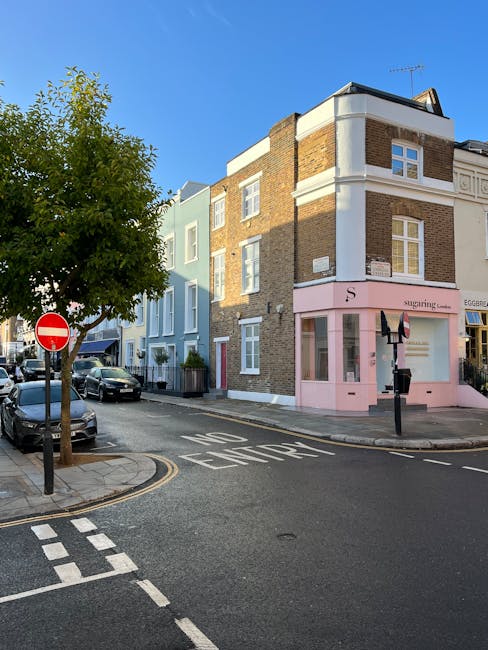 View of a narrow urban street during daytime with a four-storey corner building featuring a pastel pink shopfront on the ground floor, labeled 'sugarings', and white-framed windows on upper floors. A black bollard and a black-and-white striped motorcycle parking sign are positioned in front of the shop. To the left, parked cars line the street, with a large green tree partially obscuring a light blue building further down. The street has a 'NO ENTRY' sign and white road markings indicating a restricted zone, with a curb and tarmac surface. The scene is illuminated by natural daylight with a clear blue sky overhead, and the environment suggests a quiet local area suited for house and office removals, as part of a relocation or moving service, such as those offered by Man and Van Teddington.
