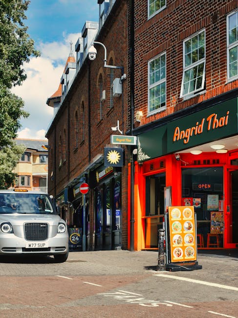 A street scene showing the exterior of a building with red brick walls and large windows, located on a busy urban high street. To the right is a Vietnamese restaurant called 'Angrid Thai' with a red storefront and an illuminated 'open' sign in the window. Next to it, a small sandwich board displays images of food offerings. On the left side of the image, a grey luxury car is parked on the street, and pedestrians are walking past shopfronts and cafes that line the pavement. Above the street, there are street signs, including a 'no entry' sign and a blue sign indicating parking restrictions, mounted on a lamppost with a white security camera attached. The background includes additional buildings with various architectural styles, and the sky above is partly cloudy with patches of blue. The scene depicts typical urban commercial activity, with visible storefronts and vehicle parking arrangements, suitable for a listing about house removals or relocation services in a busy town center.