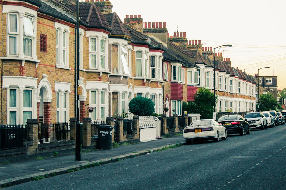 A row of traditional terraced houses in Teddington with brick facades, white bay windows, and pitched tiled roofs, lining a residential street. Several cars are parked along the curb, including a white vehicle directly in front of a house with a small front garden and black iron fence. Street lighting posts and overhead power lines are visible above the pavement. The street appears quiet with no visible people or movement, suggestive of a typical home relocation or local move setting. This scene complements house removals and furniture transport services offered by Man and Van Teddington, especially considering the narrow street that may require careful planning during the loading and unloading process for packing and moving projects.
