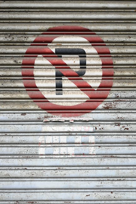 A close-up of a weathered metal shutter with horizontal slats, featuring a painted 'No Parking' symbol consisting of a black letter 'P' crossed out by a red circle and diagonal line. The shutter surface shows signs of peeling paint, rust, and dirt. The 'No Parking' sign is prominently centered, with some additional faded paint markings underneath. This image relates to house removals and moving logistics, as it highlights parking restrictions that need to be considered during home relocation or furniture transport, which [COMPANY_NAME], such as Man and Van Teddington, might need to navigate when planning removal services in the TW11 street area.