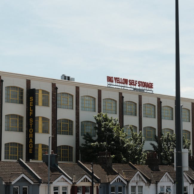 A large multi-storey building with a white facade and yellow-framed arched windows, featuring a red sign on the roof that reads 'BIG YELLOW SELF STORAGE' and a black vertical sign on the left side that states 'SELF STORAGE'. The building is situated behind a row of small, uniform residential houses with pitched roofs and brick chimneys. In the foreground, tall street lamp posts and trees partially obscure the view. The scene is captured during daylight hours with a partly cloudy sky, and the overall image conveys a commercial storage facility located within an urban area, relevant to the context of house removals, packing, and furniture transport services provided by Man and Van Teddington.