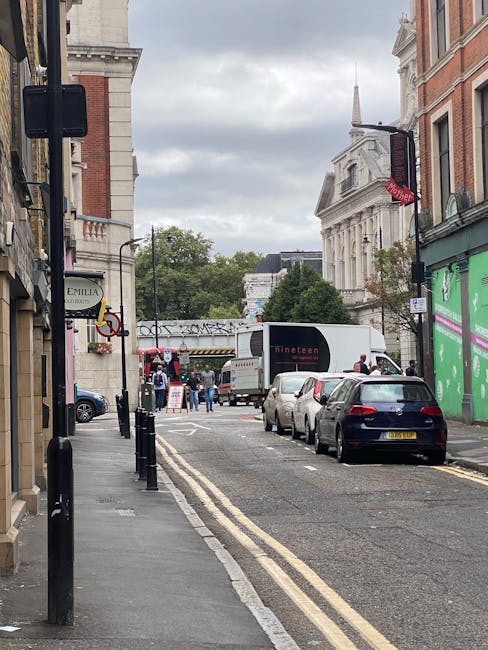 A narrow street in a city with parked cars lining both sides, including a dark blue vehicle closest to the camera and several others further along. On the left, there is a row of buildings with shop signs, one reading 'Emilia' and another partially visible. Street bollards are positioned along the pavement, and the road features double yellow lines indicating parking restrictions. In the distance, there are people gathered near a white moving truck with black branding, which is parked partially on the pavement, suggesting an ongoing home relocation or furniture transport process. The background includes a mix of historic and modern architecture with trees and a cloudy sky overhead. This scene reflects typical urban logistics, where [COMPANY_NAME] might be involved in removals or house moving services in Teddington, focused on easing access problems during relocation.